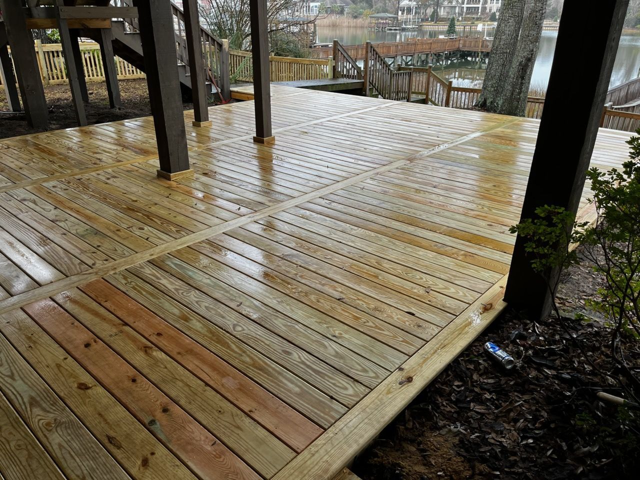 A ground-level wooden deck with wet planks, dark support posts, and a view of a lake and backyard in the background.