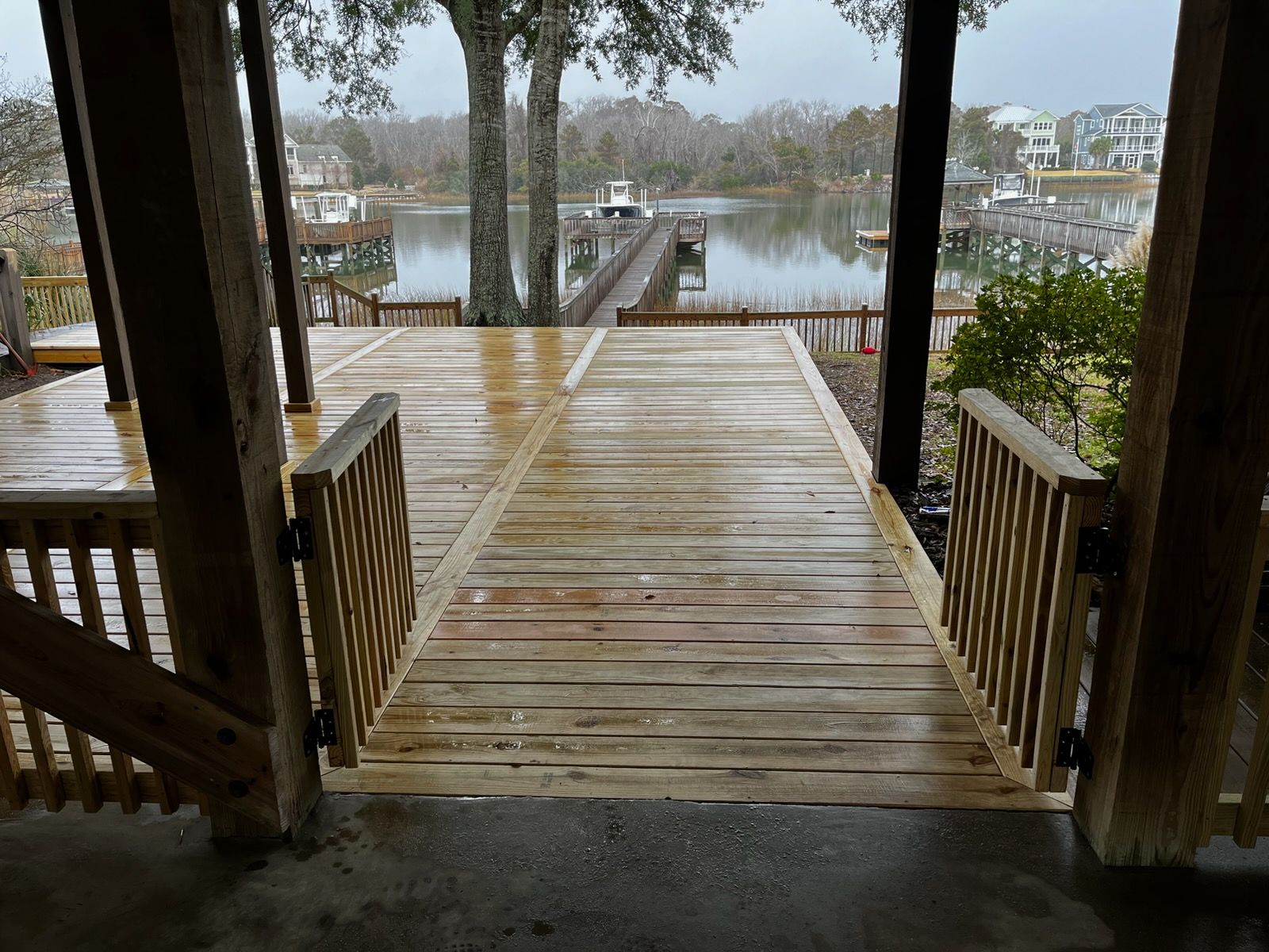 A wooden deck gate opens to a pathway leading toward a dock and water on a cloudy day.