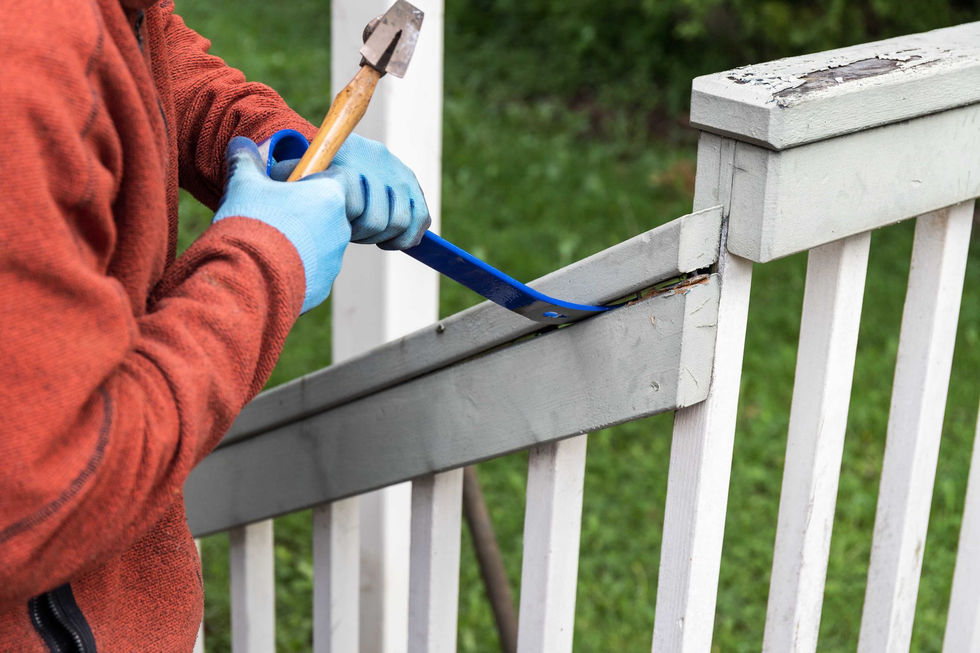 A person wearing blue gloves uses a blue crowbar to pry a wooden railing off a painted white fence outdoors.