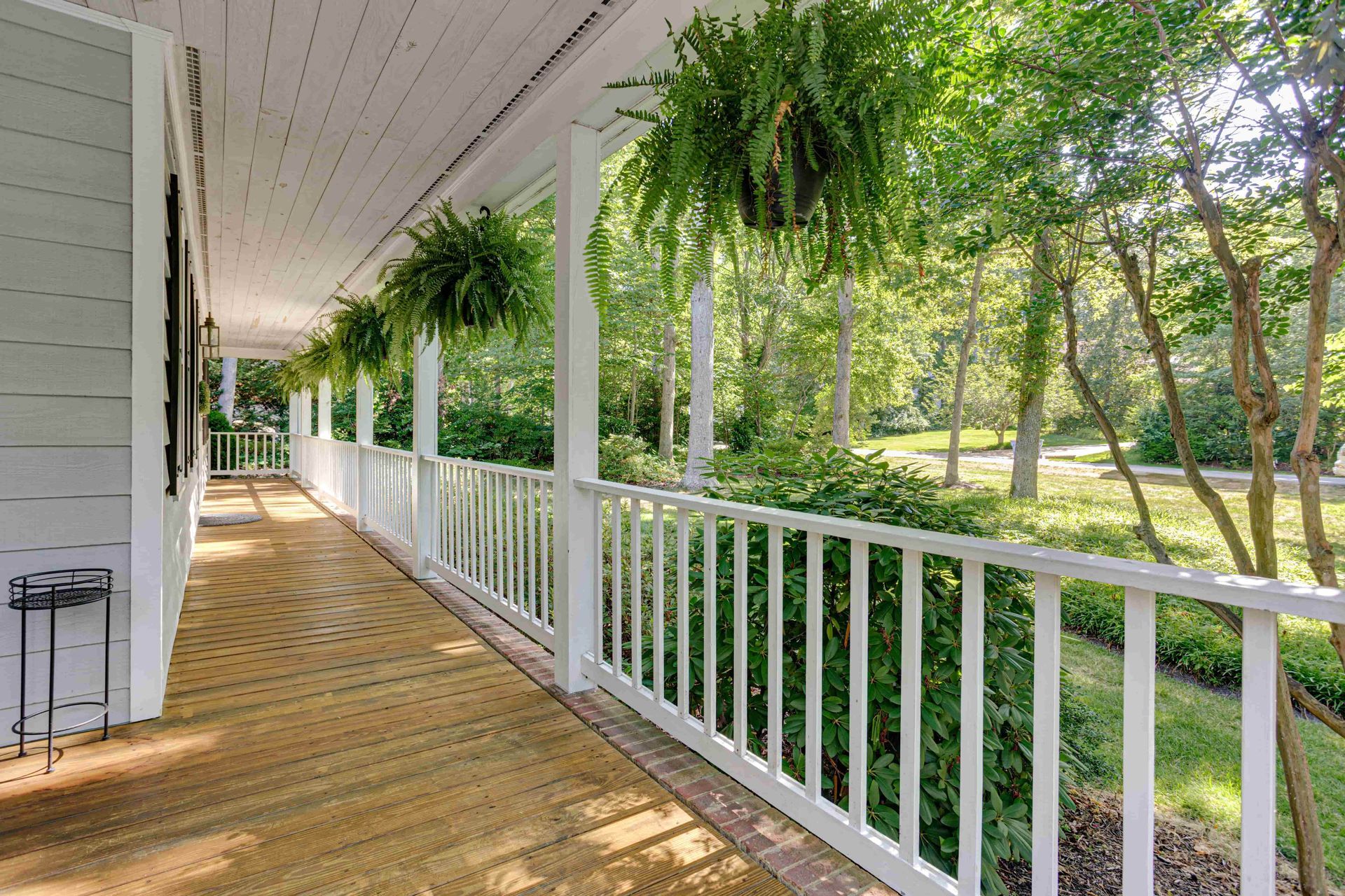A long, light gray porch with wooden flooring, white railings, and hanging ferns, overlooking a sunlit yard and trees.
