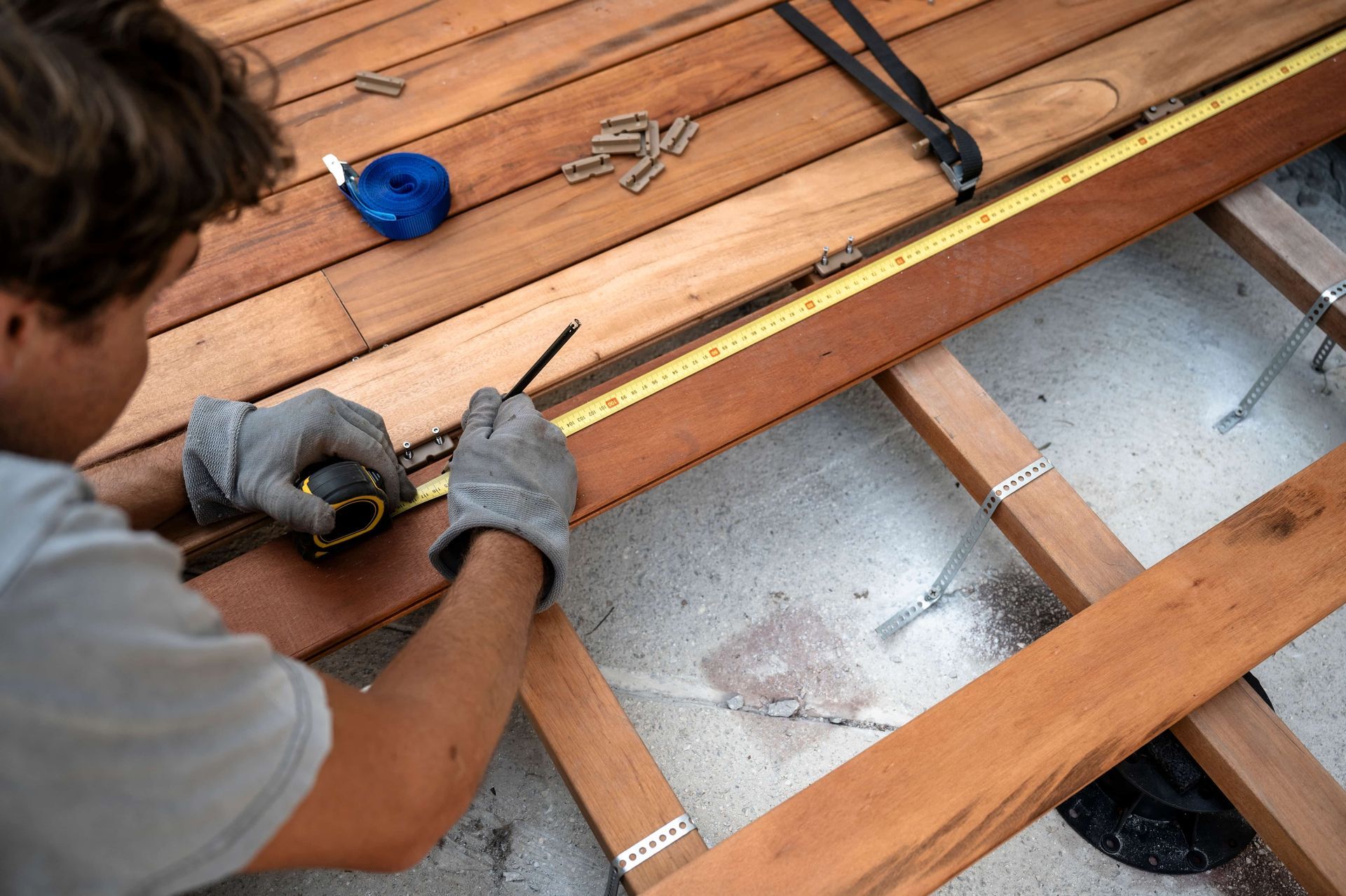 A person wearing gray work gloves uses a pencil and tape measure to mark wood planks on an outdoor deck frame.