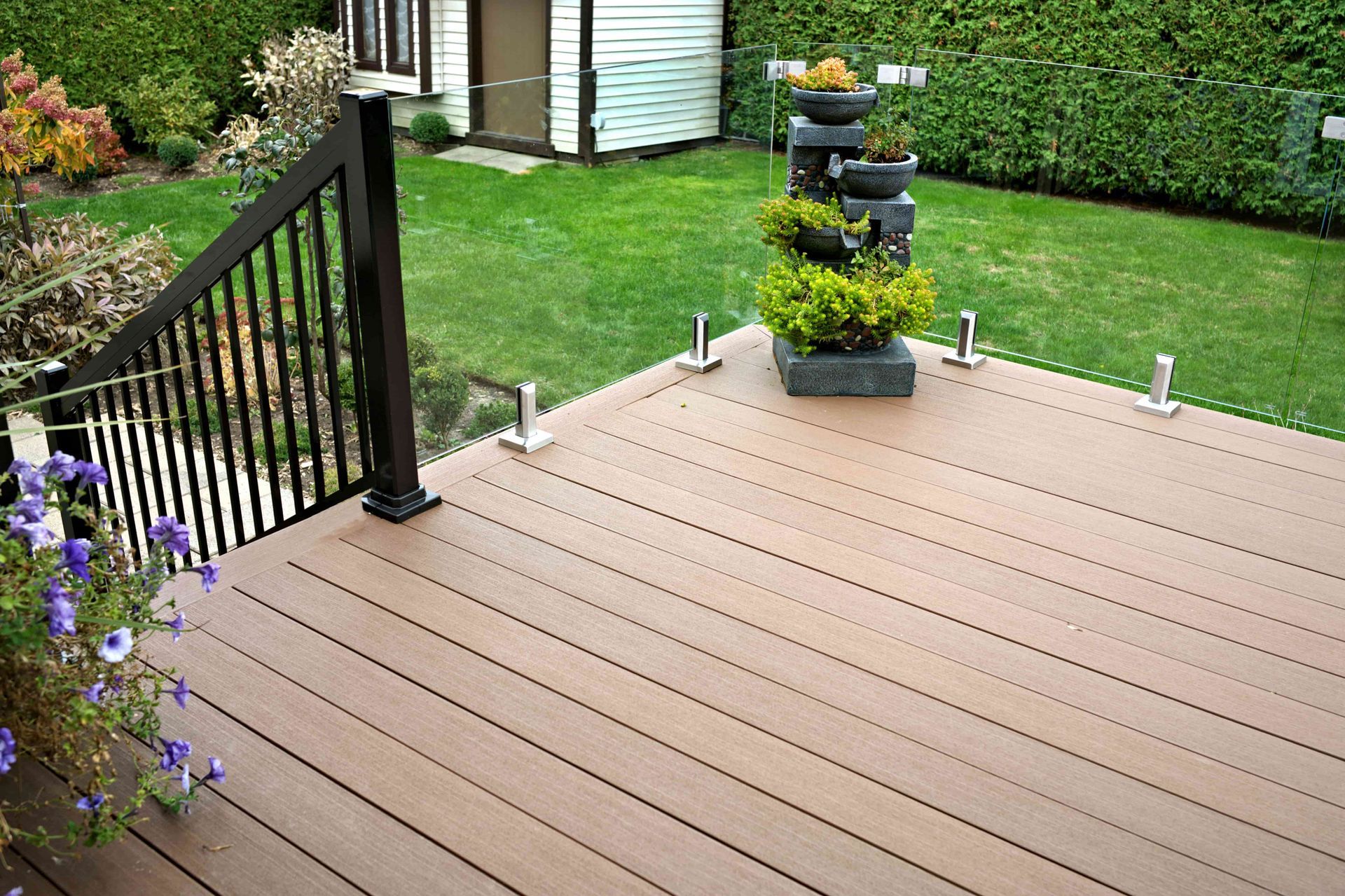 A composite wood deck features a black railing, glass panel borders, and a tiered stone fountain in a lush green backyard.