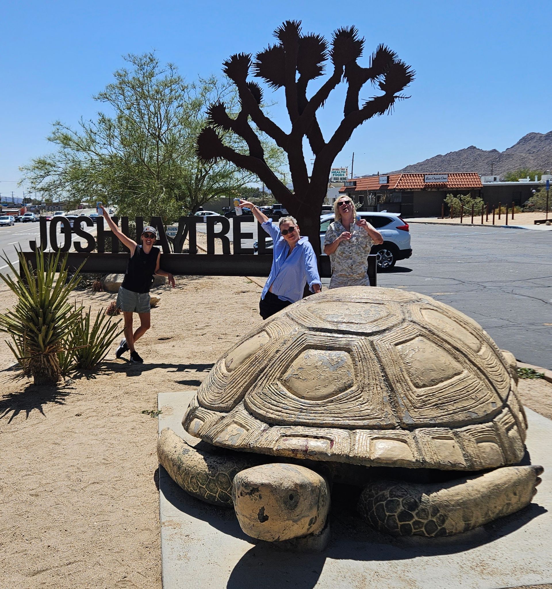 Three people pose near a large tortoise statue and a "Joshua Tree" sign. Desert setting, sunny day.