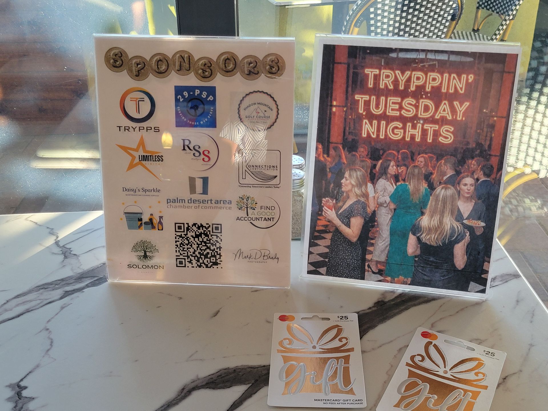 Two signs and gift cards on a table. One sign lists sponsors, the other says "Tryppin' Tuesday Nights" above a group photo.
