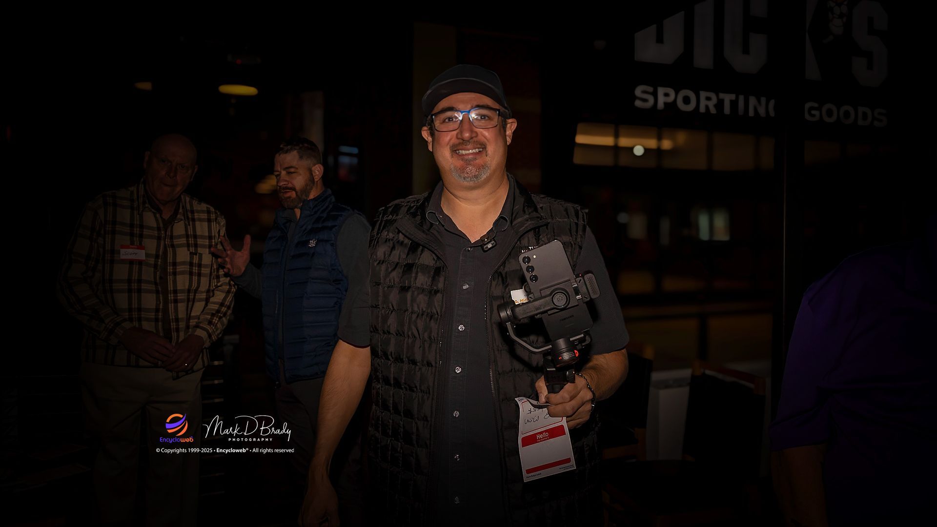 Man in black vest and hat holding a camera, smiling outside a sporting goods store.