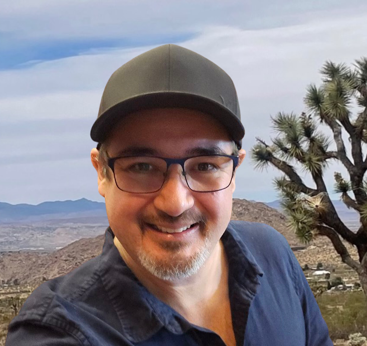 Smiling man in glasses and a black cap outdoors with mountains and desert in the background mike the driver