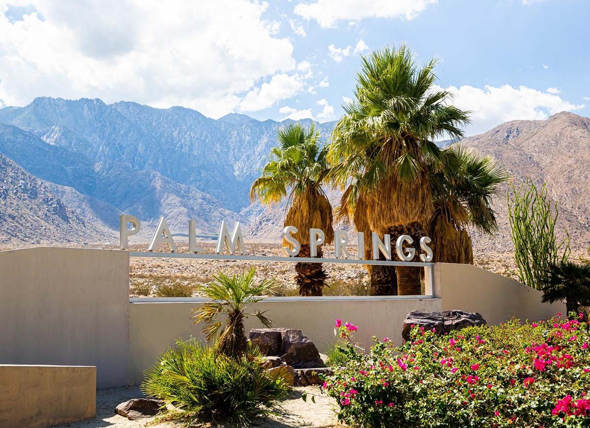 A sign for palm springs is surrounded by palm trees and flowers with mountains in the background.