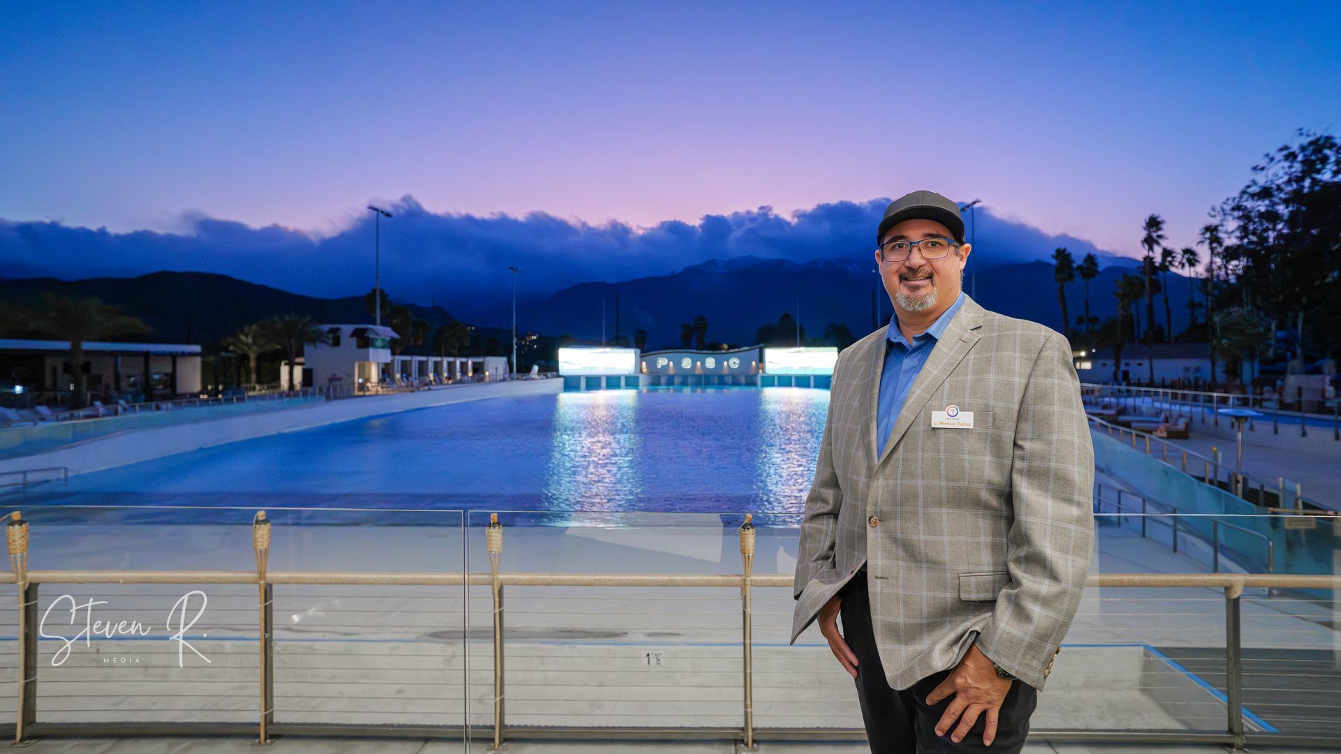 Man standing near a wave pool at dusk, wearing a blazer and hat; mountains in the background.