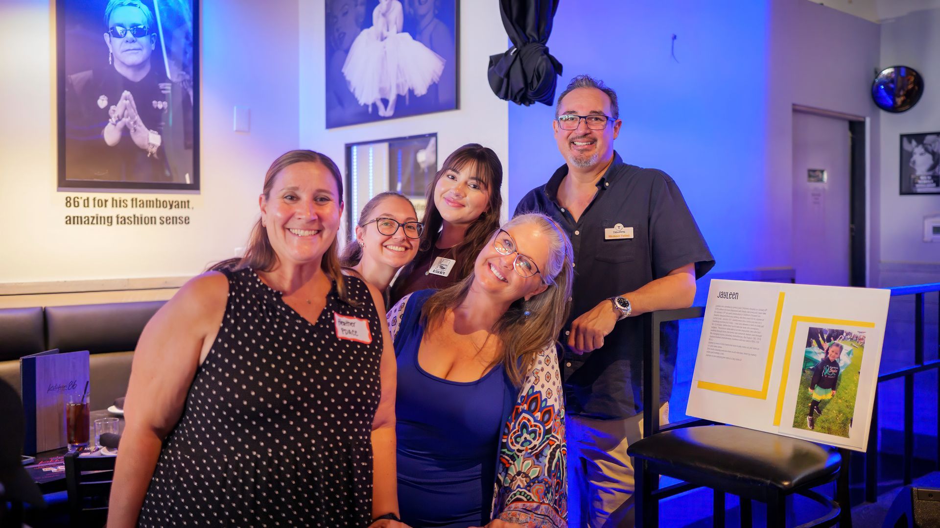 Group of five people smiling at a bar, with photos on the wall and a memory board.