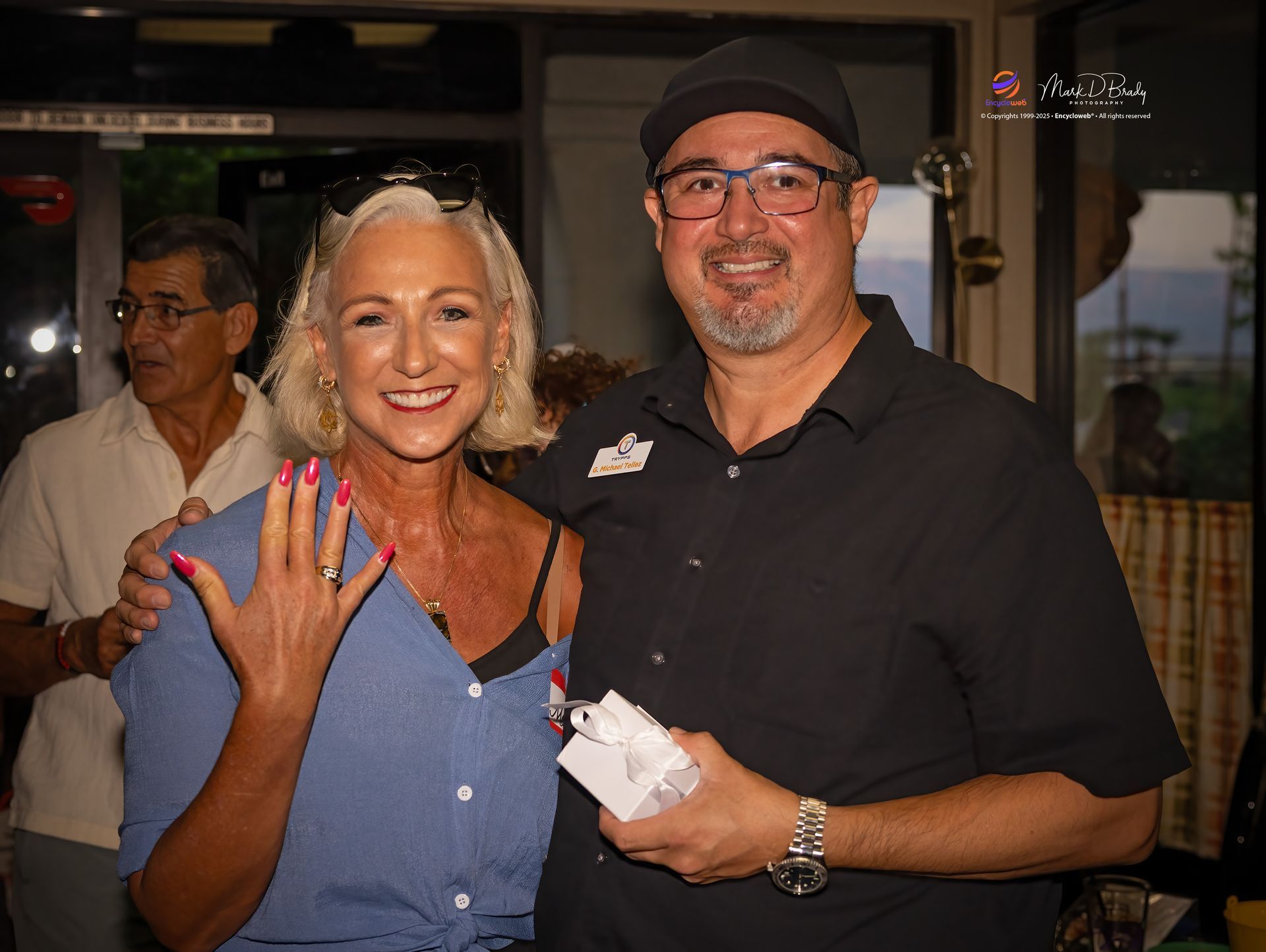 Woman shows ring, smiles; man holds ring box, smiles. Indoor setting.