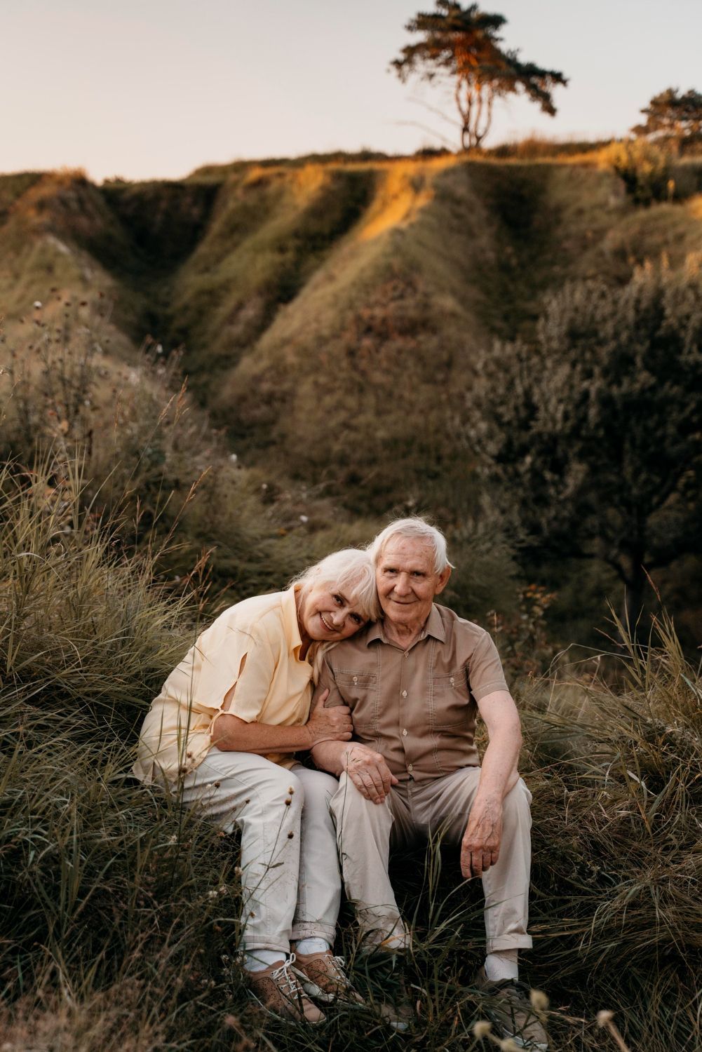 An elderly couple is sitting in the grass with a tree in the background.