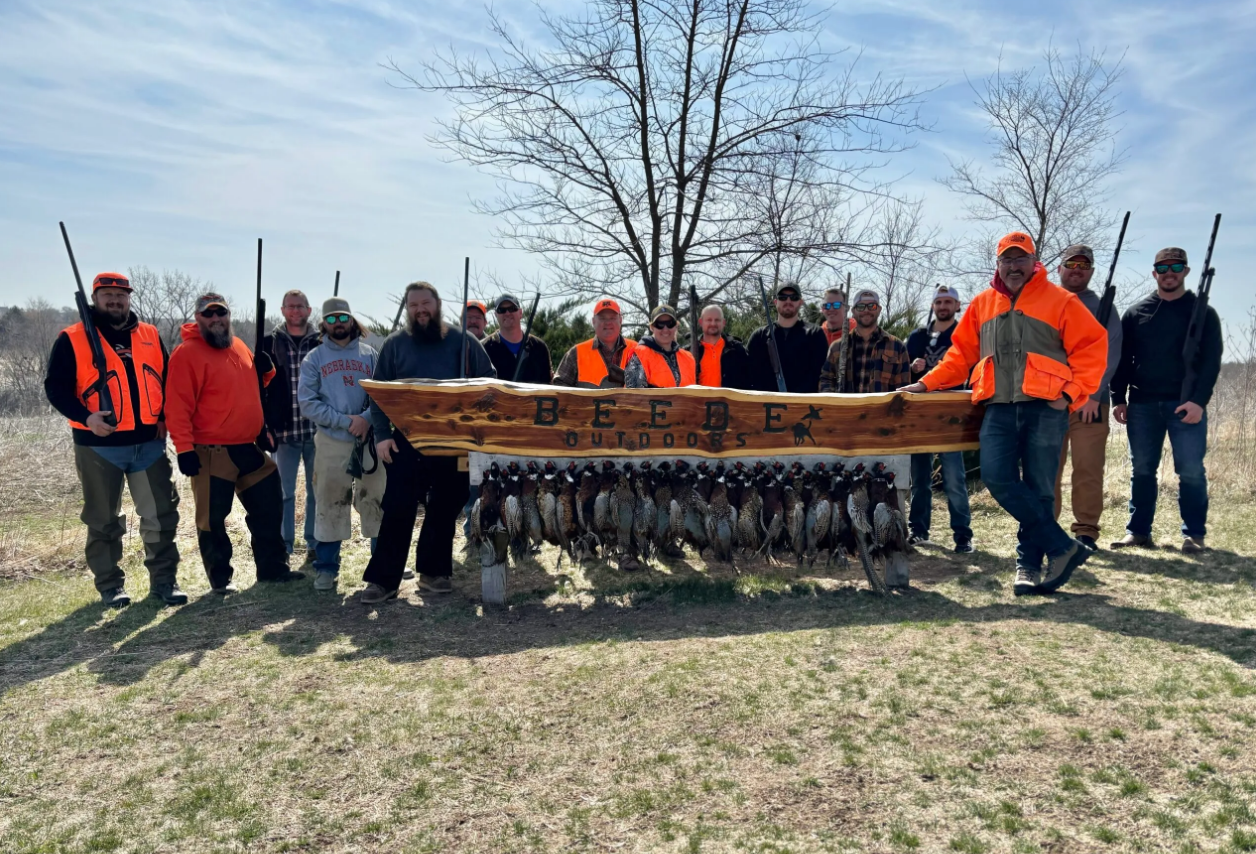 Group of hunters with shotguns pose with harvested game birds in a field; they are wearing orange vests.