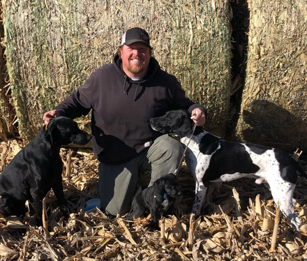 Man kneels with four dogs in a field. Dogs are black, black/white, and two small black ones.
