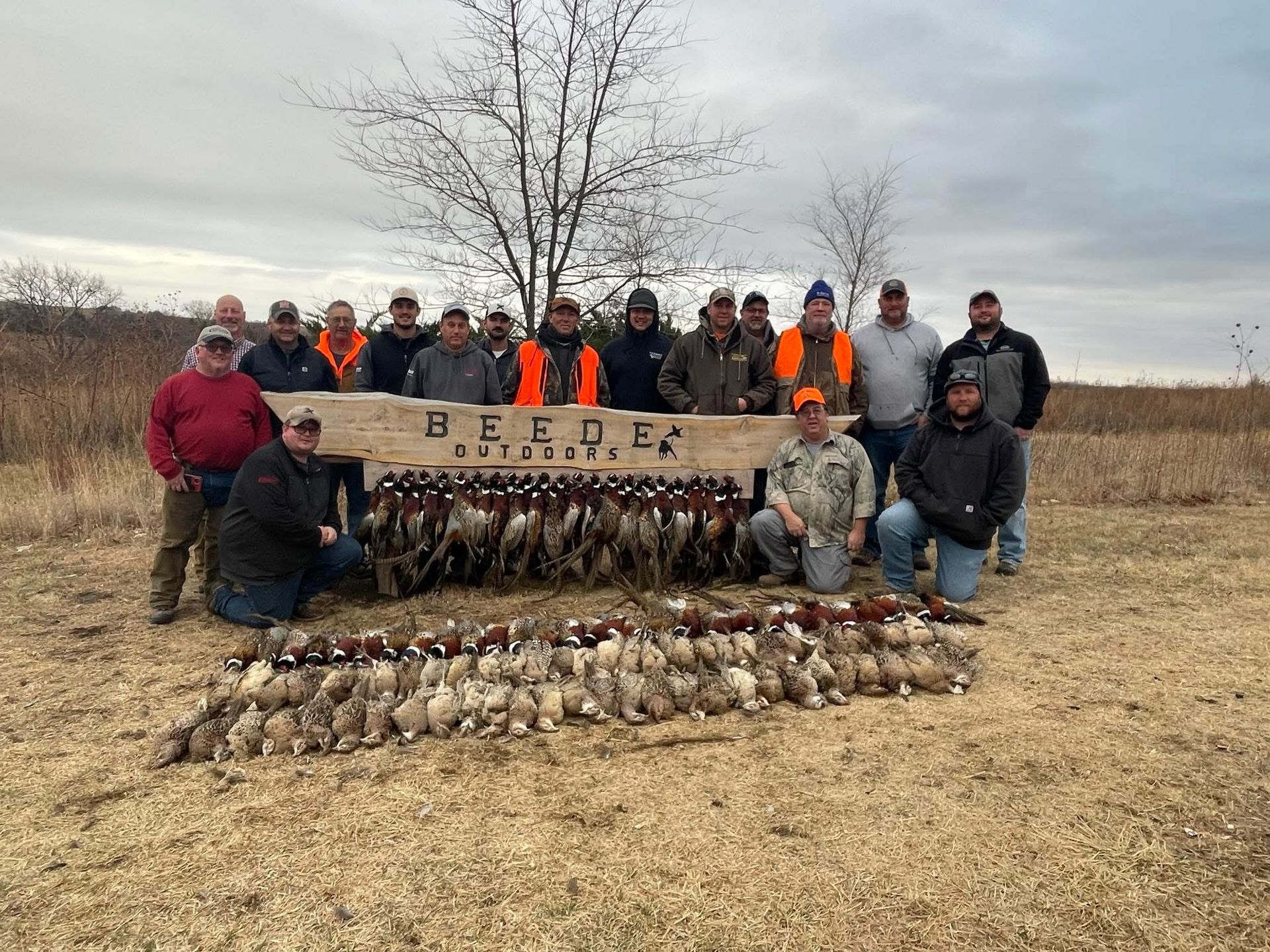 Group of men posing with hunted pheasants in a field. Some are wearing orange vests, autumn setting.