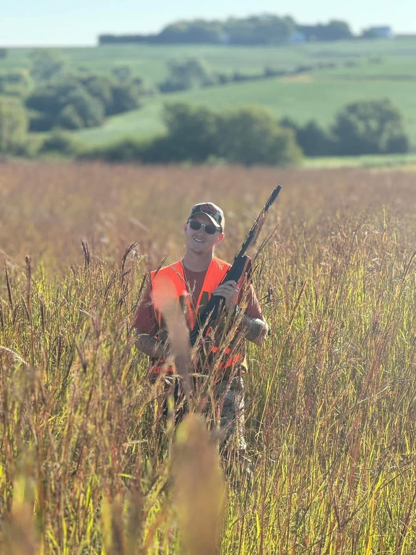 Hunter in orange vest, holding a shotgun, standing in tall grass in a field.