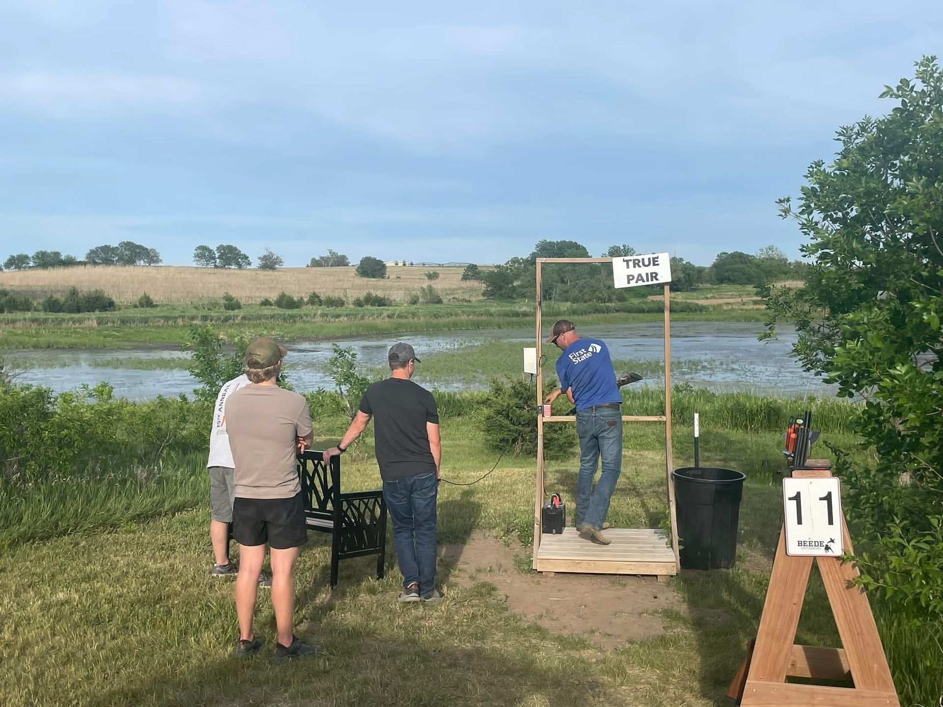 People shooting clay pigeons at an outdoor range. Shooter takes aim, others watch. Water and fields in the background.