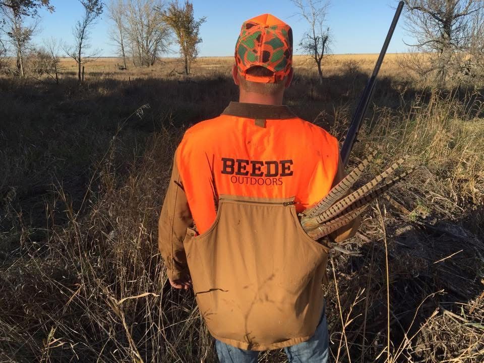 Hunter in orange vest with pheasant feathers, standing in field.