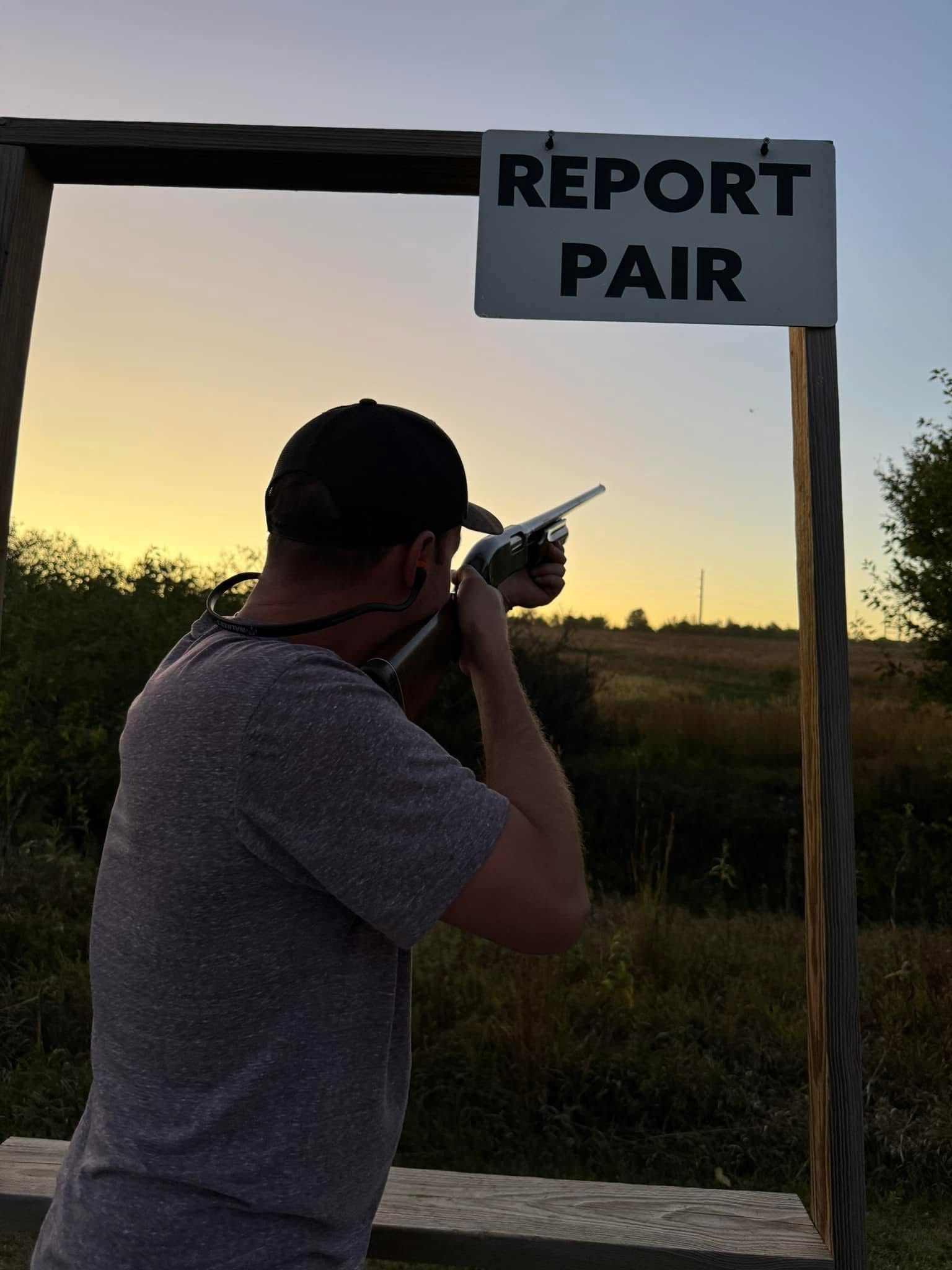 Man aiming a firearm at a target outdoors, under a sign that reads 