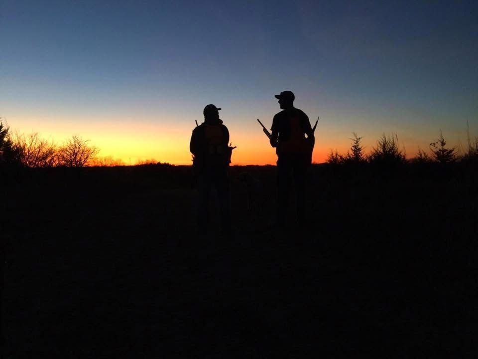 Two hunters silhouetted against a sunset, holding rifles in a field.