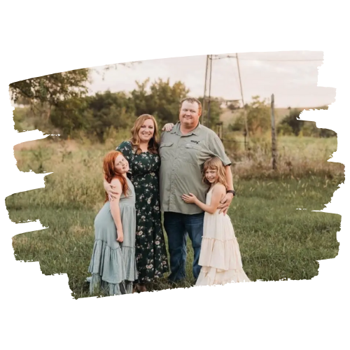 Family of four in field posing, smiling. Mother in floral dress, father in green shirt, two girls. Windmill in background.