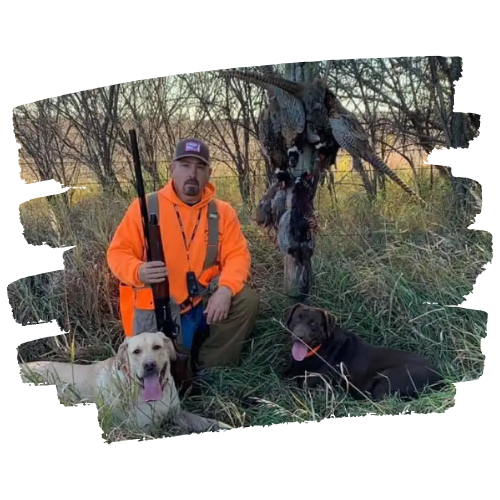 Hunter in orange, with two dogs, holding a shotgun next to harvested game.