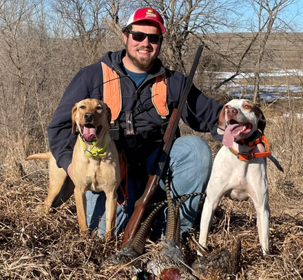 Man kneels with two hunting dogs, smiling. Brown shotgun, dead bird. Outdoors, sunny day.