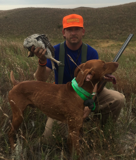 Hunter kneeling in field with a bird and dog, wearing an orange hat. Dog is brown and excited.