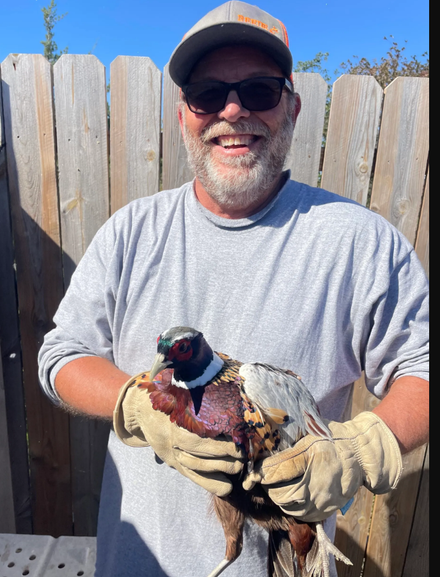 Man in cap and sunglasses smiles while holding a pheasant, standing outdoors by a wooden fence.