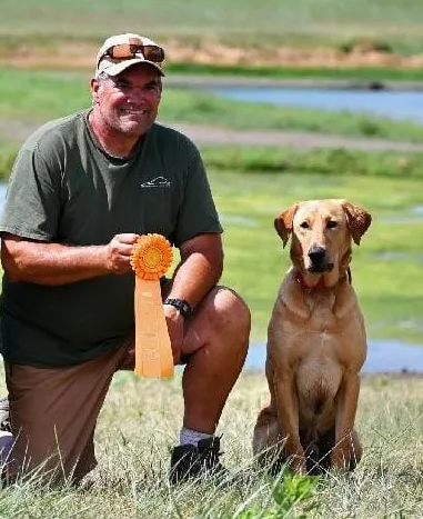 Man kneeling with a yellow Labrador, holding a ribbon outdoors. Green grass and water in the background.