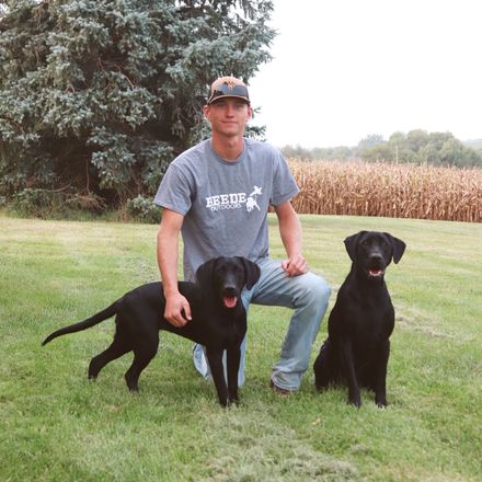 Hunter kneels in snow with two dogs and hunted birds. Wearing orange vest, hat. Snowy outdoor setting.