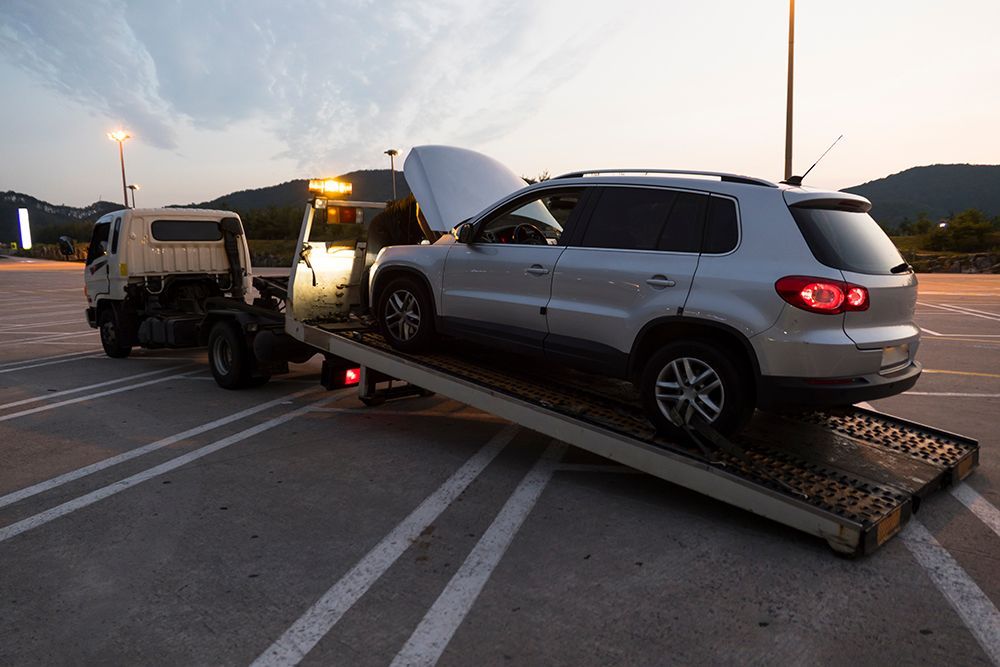 Silver SUV on a tow truck ramp in a parking lot at dusk, with the hood open.