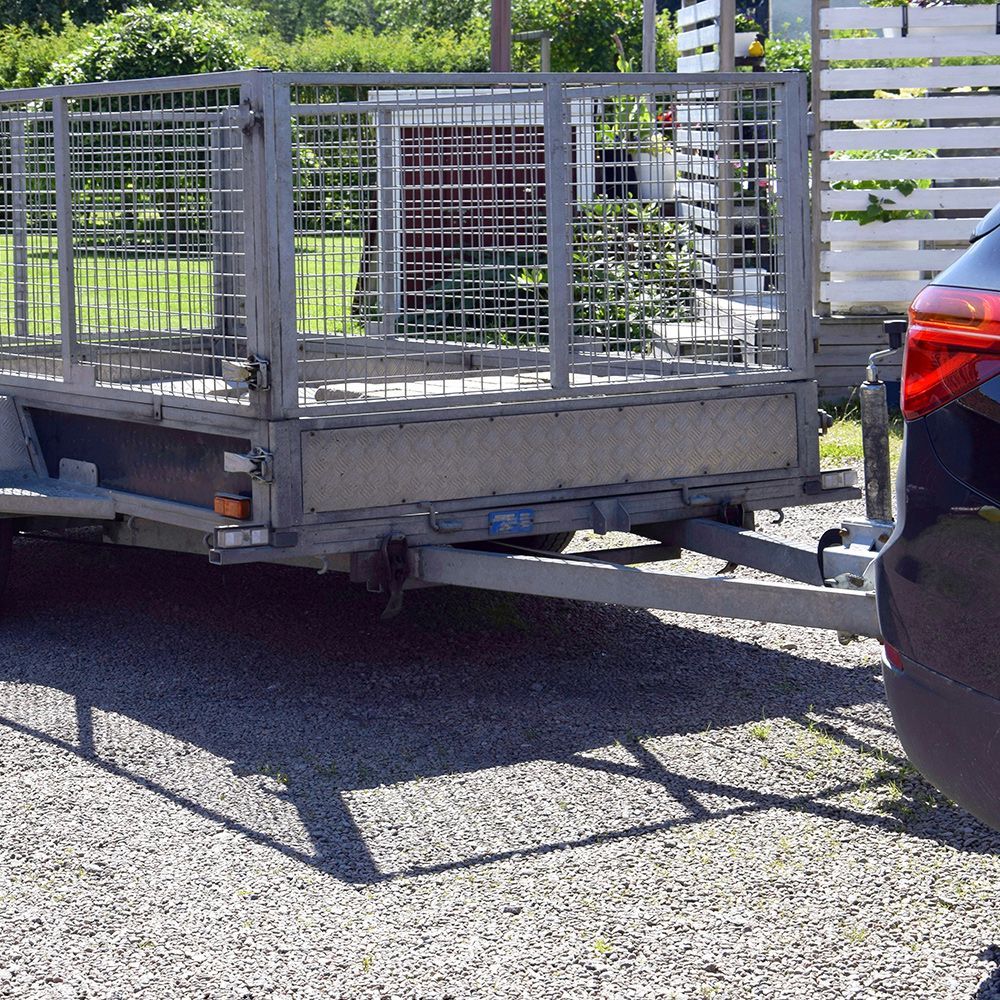 Gray utility trailer with metal cage hitched to a black car on a gravel driveway.