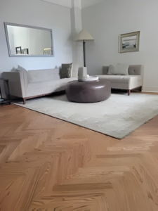 Living room with beige sectional sofa, brown ottoman, light rug, and herringbone wood floor.