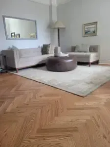 Living room with beige sectional sofa, brown ottoman, light rug, and herringbone wood floor.