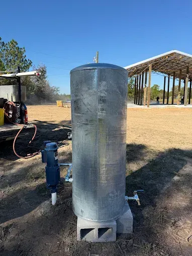 A tall, galvanized steel well pressure tank sits on concrete blocks next to a pump in a sunny, rural construction site.