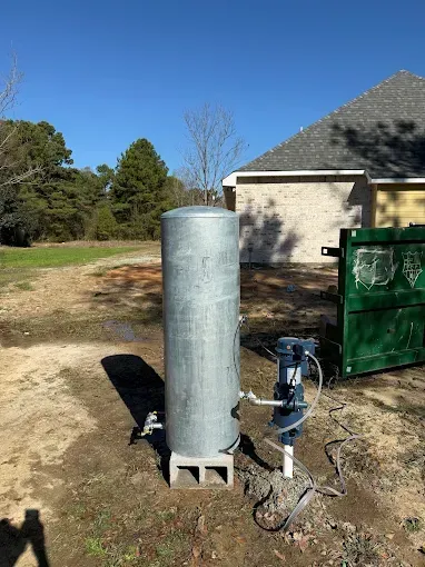 A tall, silver pressure tank on concrete blocks next to a water pump outside a house on a sunny day.