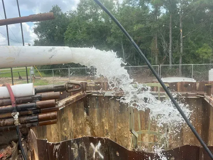 Water gushing from a white industrial pipe into an excavated construction trench held by metal sheet piling.