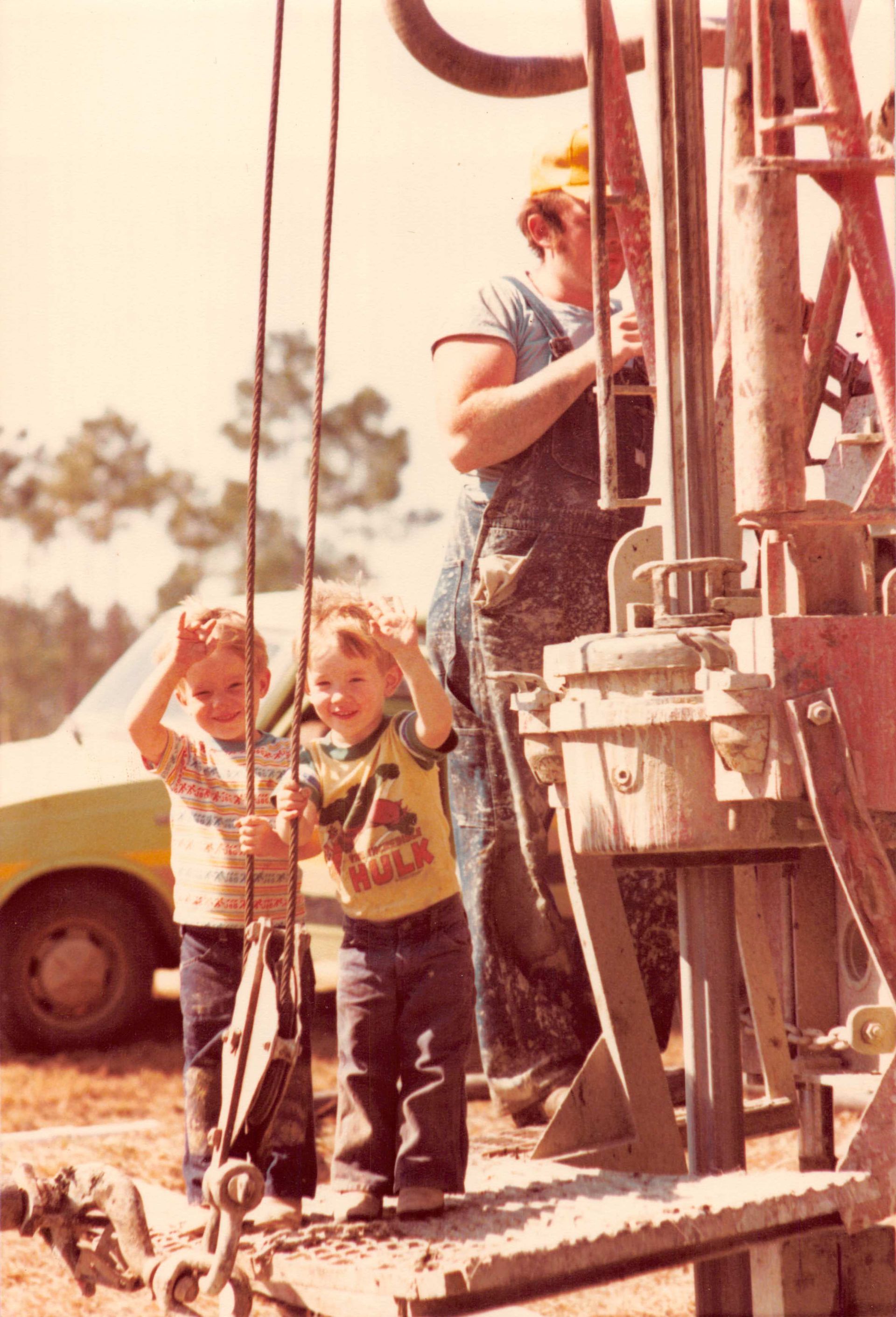 Two children stand on a drill rig platform smiling and making bunny ears with their hands, while an adult operates the rig.