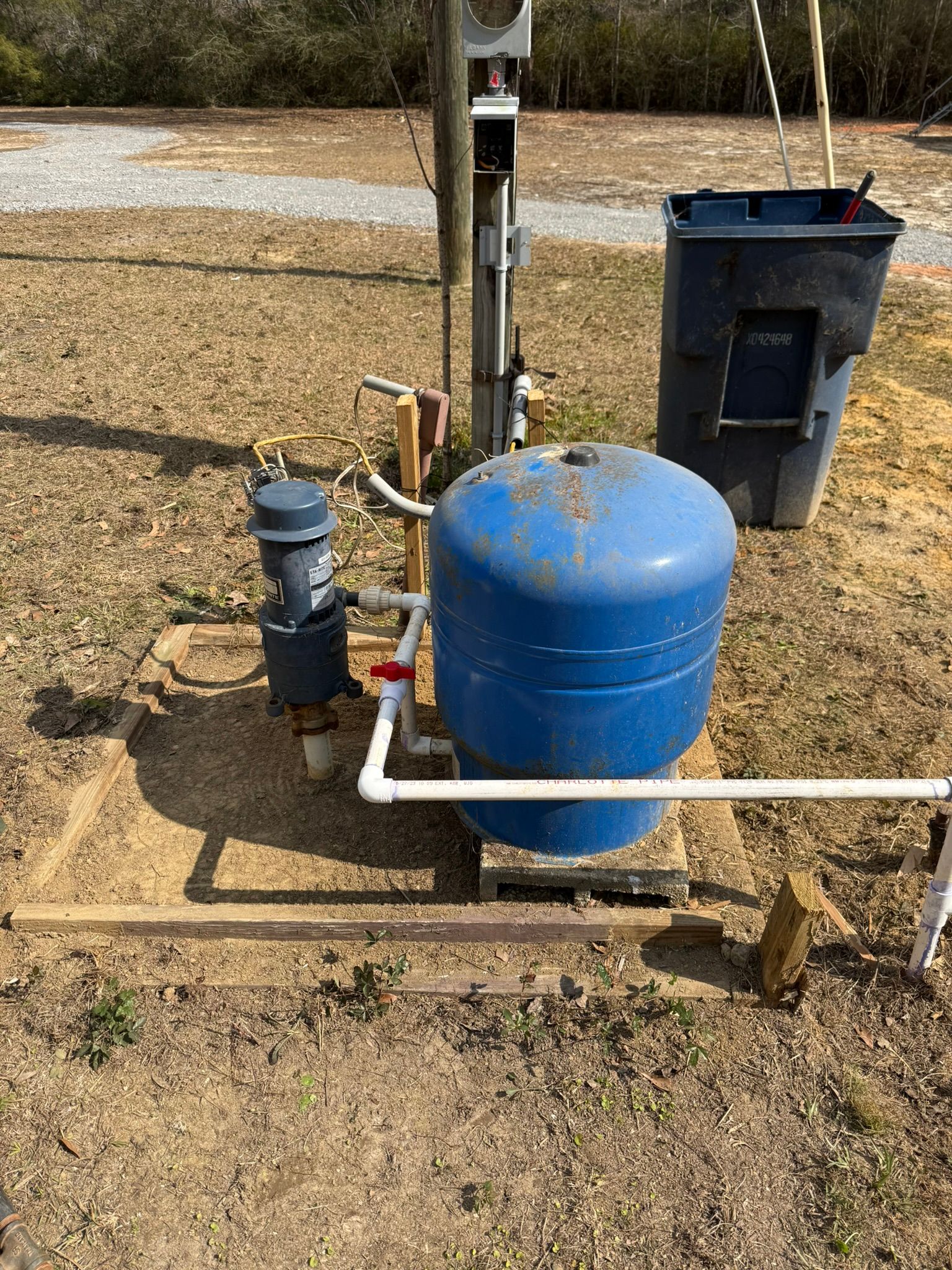A blue water pressure tank and pump system installed outdoors on a concrete pad with a utility pole and bin nearby.