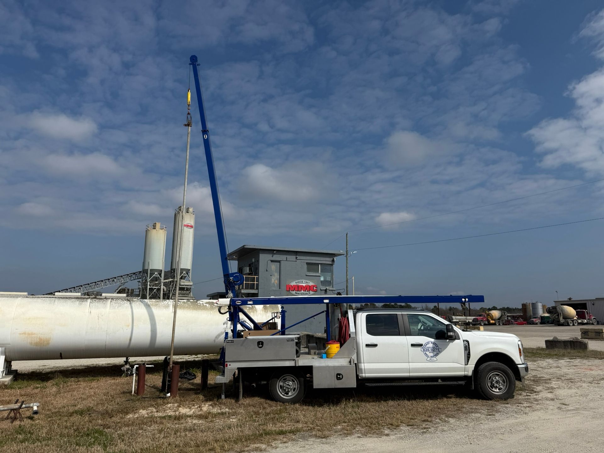 A white service truck with a tall, blue extended crane arm parked on a gravel lot near industrial storage tanks.