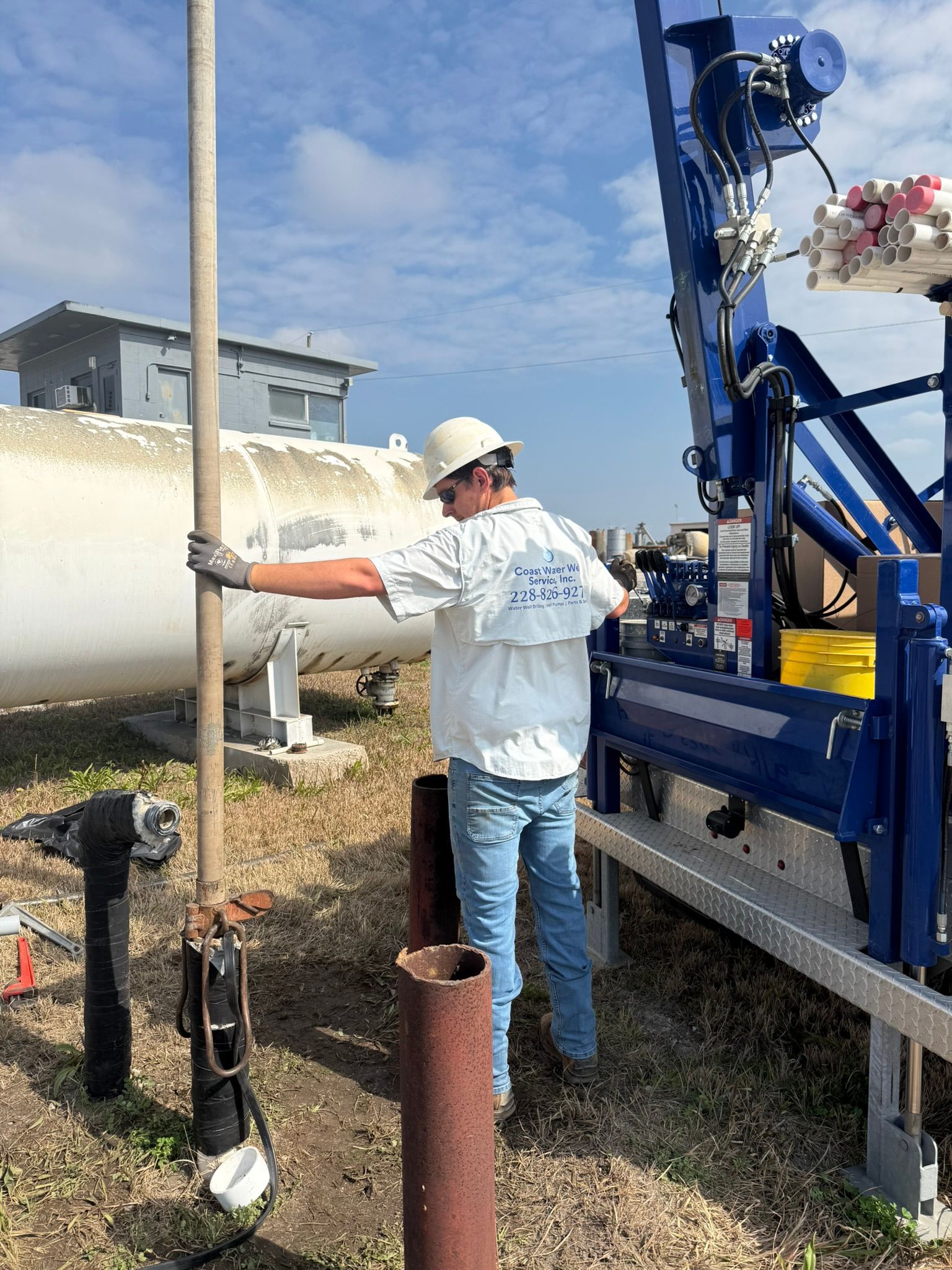 A person in a hard hat and work clothes holds a long metal pole near a drilling rig at an industrial site.