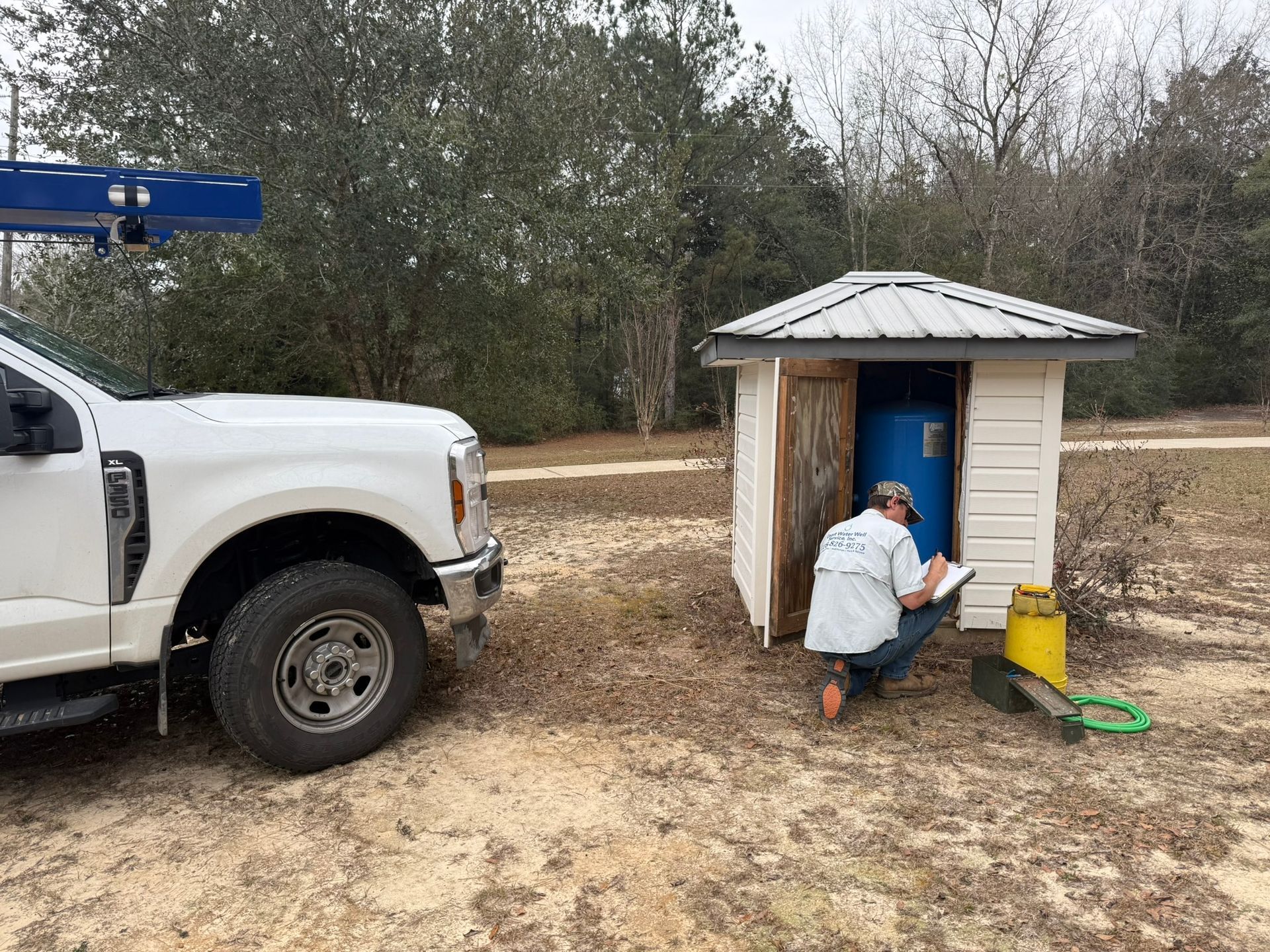 A technician wearing a white shirt kneels outside a small shed, documenting work on a blue water tank near a white truck.