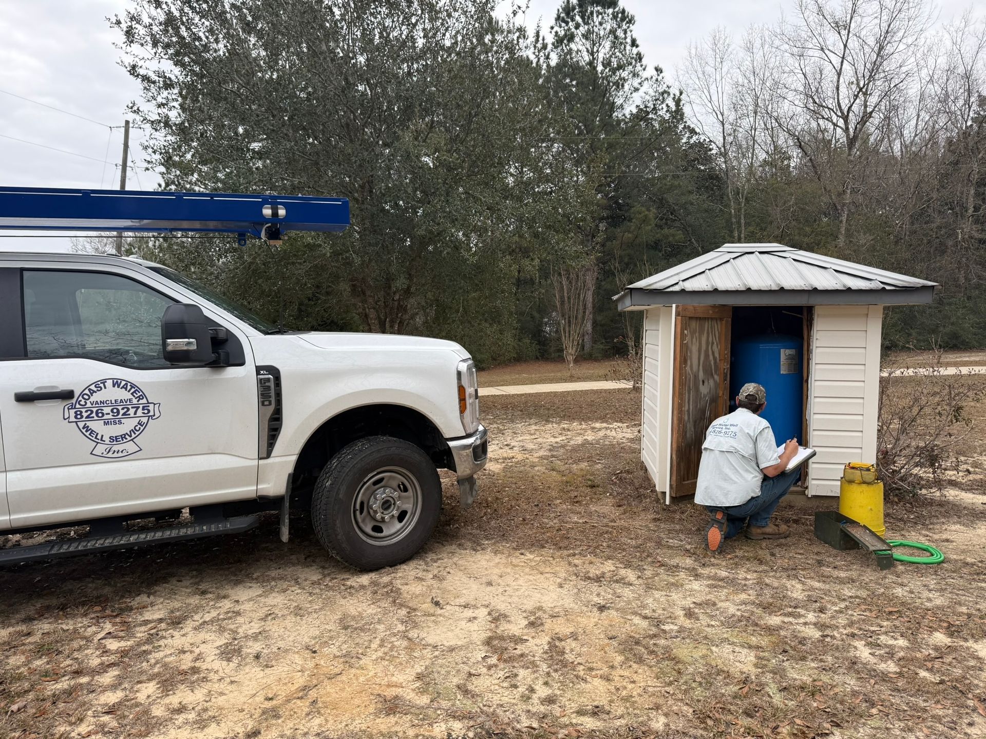 A technician sits by an open utility shed checking a blue tank, with a work truck parked nearby on a dirt lot.