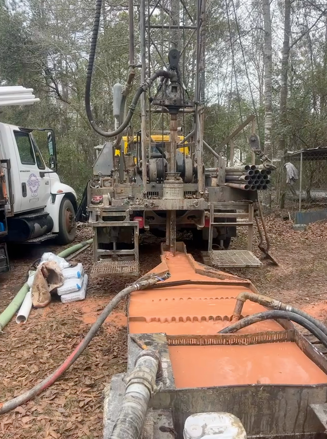 A yellow drilling rig in a wooded area, pumping orange-tinted liquid into a metal trough.