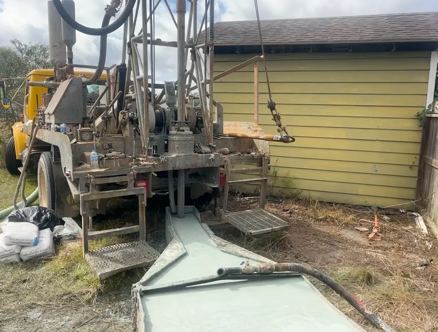 A yellow drill rig operates on a dirt lot, pumping light-colored slurry into a metal trough beside a pale yellow building.