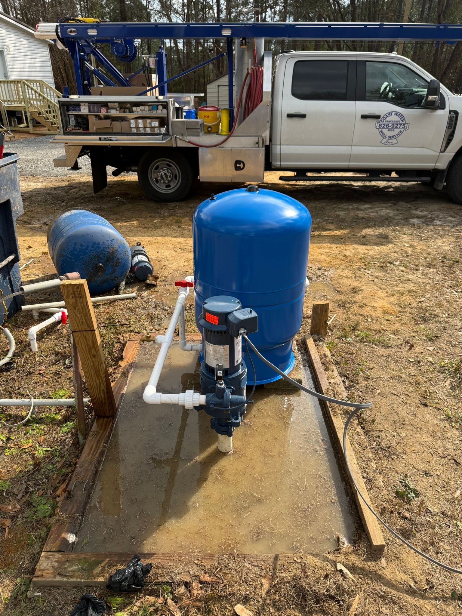 A blue water pressure tank and pump system installed on a concrete pad outdoors near a service truck.