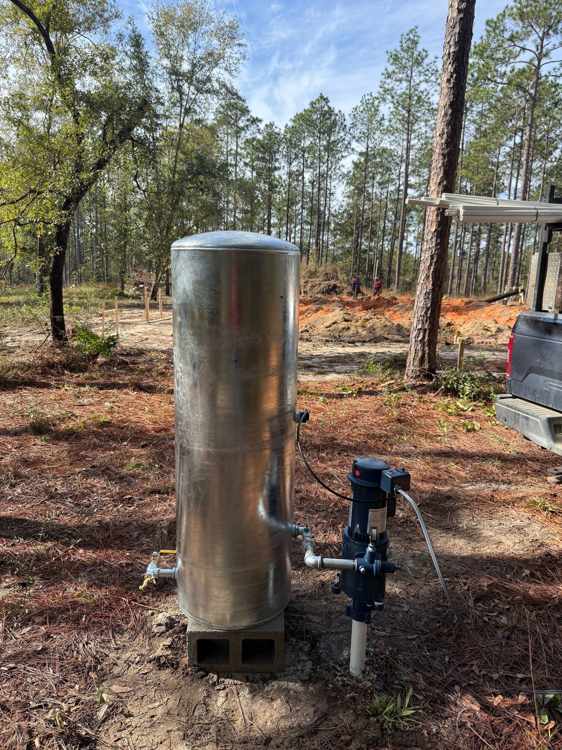 A large, vertical galvanized water pressure tank sitting on concrete blocks in a wooded, outdoor setting.