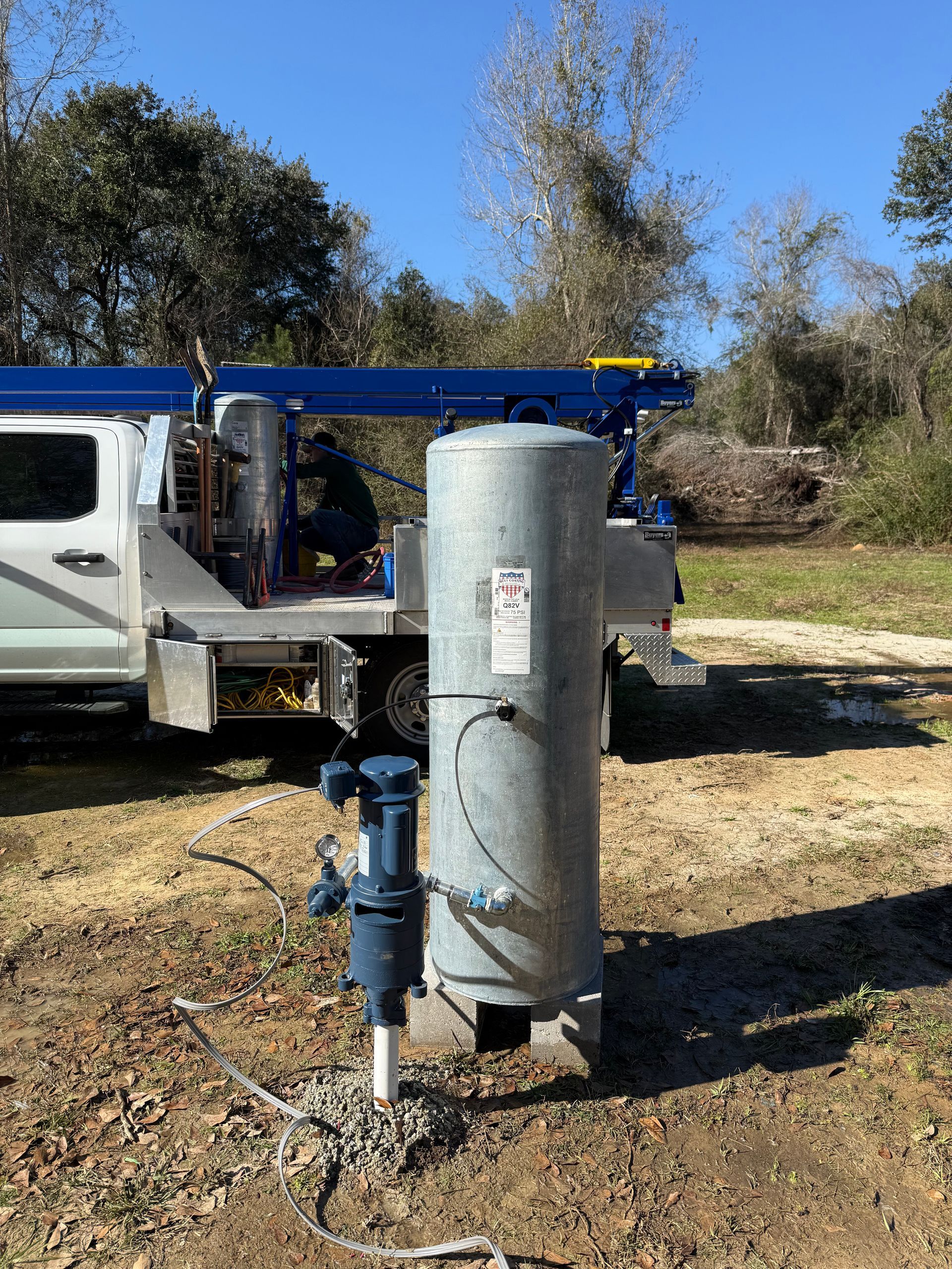 A blue water pressure tank and pump system installed on a concrete pad outdoors near a service truck.