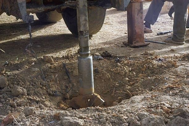 A drilling rig operates in a wooded area, discharging orange mud into a collection tank in the foreground.