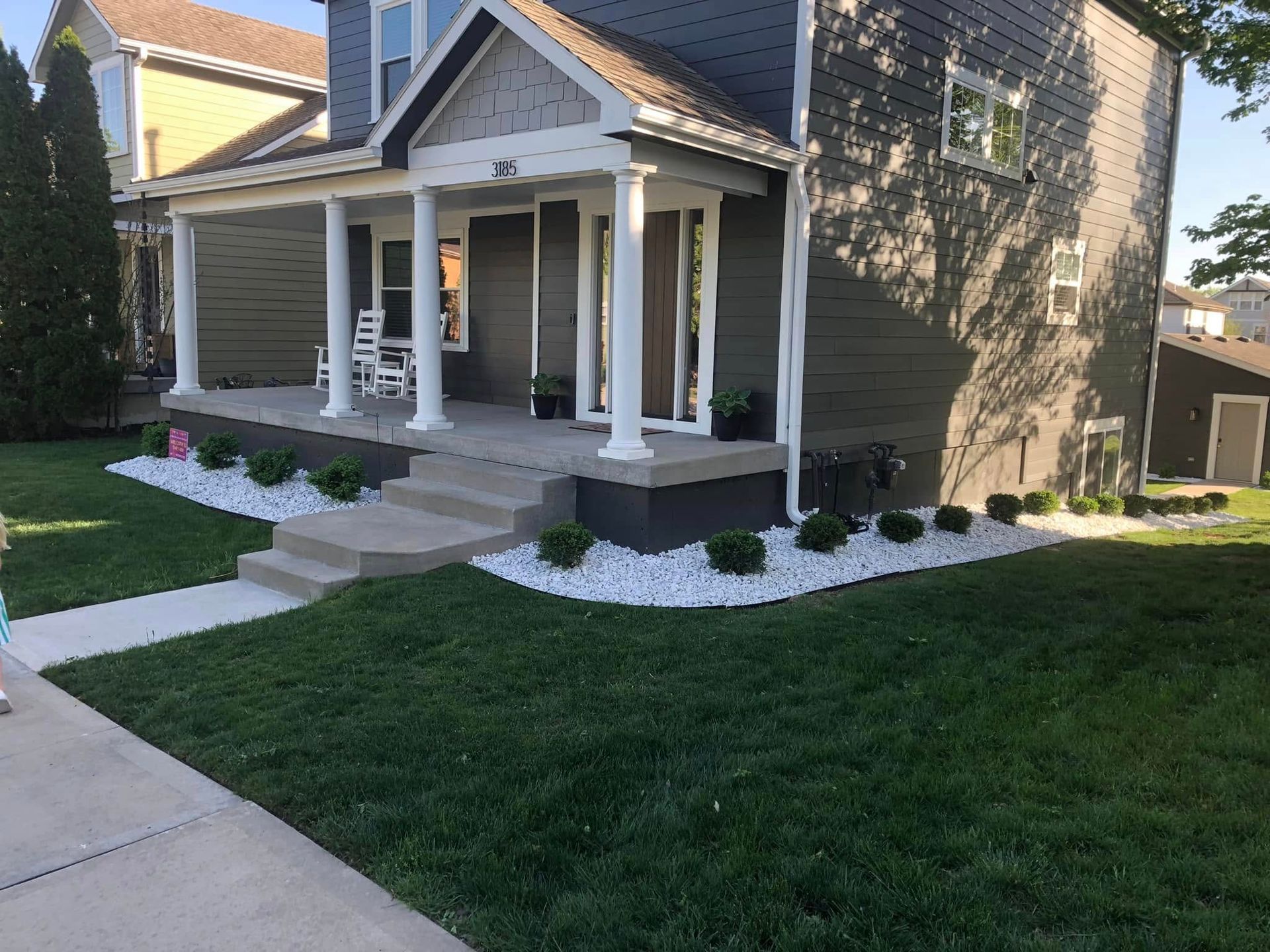 A house with dark gray siding, a white front porch with pillars, white landscaping stones, and a green lawn.